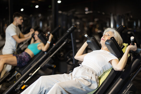 Positive elderly woman in great shape using hack squat machine in busy gym, showing healthy aging, mobility, and active lifestyle