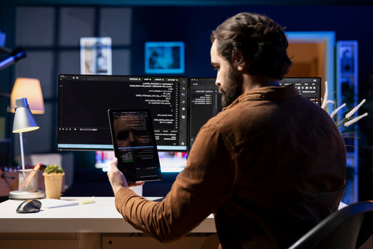 Programmer doing software debugging using tablet, checking glitches in lines of code displayed on computer monitors. Man working from home fixing coding issues using portable digital device.