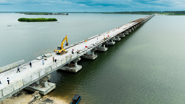 Aerial view of a bridge under construction with a yellow excavator and workers on the concrete deck over calm water with green islands in Rivers State, Nigeria.