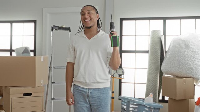 Man holds drill in hand and smiles among stacked moving boxes and rolled rug in a bright building foyer; new home pride.
