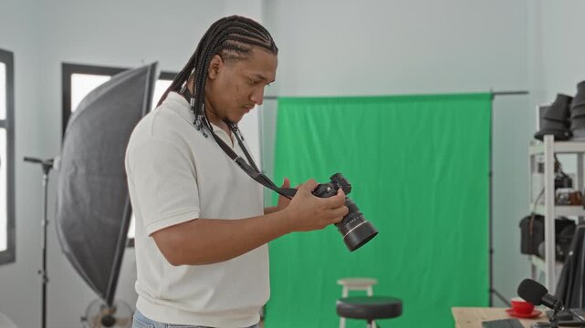 Man photographer in white shirt checks camera screen and adjusts lens near green screen and studio lights in studio; concentration preparation.