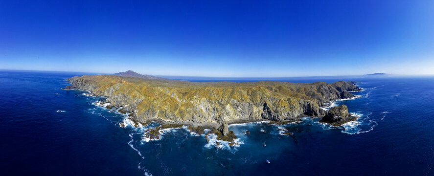 Aerial view of the rugged cliffs and rocky coastline of Magdalena Bay with waves crashing against the shore under a clear blue sky in Mexico.