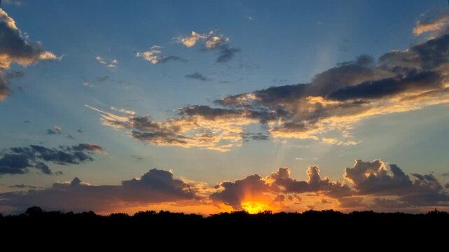 4K timelapse video capturing a golden sunrise over a dark forest silhouette. Dramatic sunbeams break through moving fluffy clouds in the blue sky, creating a peaceful and majestic morning atmosphere.