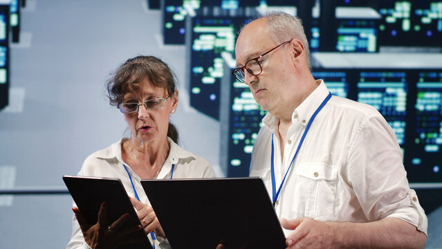 Data storage company executives looking around data center, using laptop and tablet to crosscheck contingency plan and assess high-tech workspace units in need of fixing, preventing liabilities.