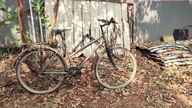 Bogor, Indonesia - June 16, 2026: Rusted bicycle leans against weathered wooden fence. Dried leaves blanket the ground around it. Sunlight filters through branches above. The scene evokes quiet abando