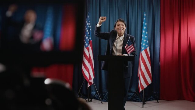 Camera filming young Hispanic woman politician standing at podium raising fist in victory during speech with USA flags in background. Clip is ideal for political campaigns and election news