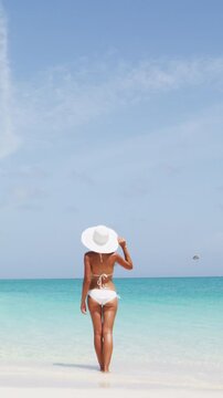 Beach bikini woman standing by ocean enjoying paradise beach resort. Young lady wearing white bikini and and elegant sun hat during summer vacation. Idyllic beach view sunny day. Vertical Video.