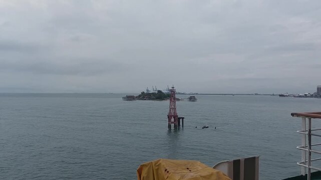 Calm ocean viewed from the deck of a moving ship near Makassar, Indonesia. Static camera captures a cloudy afternoon seascape with a navigation beacon, small island, and distant port facilities.