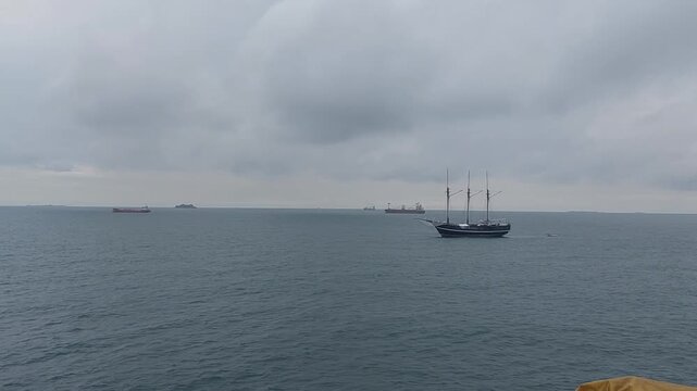 Wide seascape with anchored ships and a traditional three-masted sailing vessel on calm ocean waters under an overcast sky. Maritime transportation scene featuring cargo ships on the horizon.