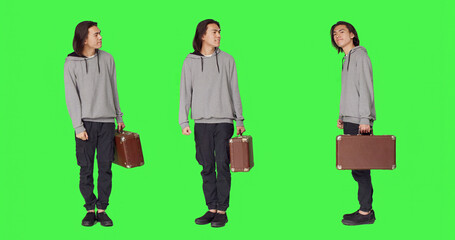 Obraz premium Person waiting impatiently in studio, looking around and checking hour on wristwatch, young adult carrying retro briefcase standing over full body green screen backdrop, acting impatient.