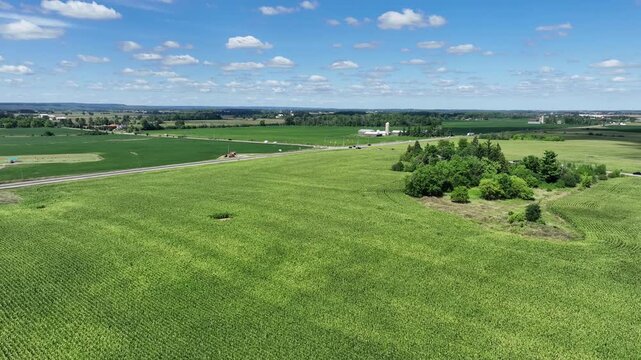 Low-level aerial drone flyover of a vibrant green corn field beside a rural road intersection, with farm buildings, silos, and open countryside stretching to the horizon.