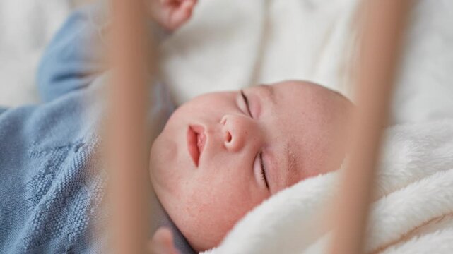 Newborn boy lying in a wooden nursery crib, mouth slightly open, eyes closed and hand lifted over white blanket; peaceful rest.