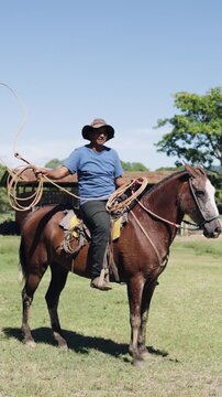 Hispanic man wearing a hat and sitting on his horse while practicing with a lasso. Professional cowboy working on a sunny day at the ranch