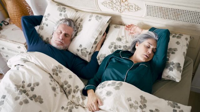 Annoyed senior husband suffering from insomnia while his wife snores loudly in bed. Man covering ears with pillow, struggling to sleep