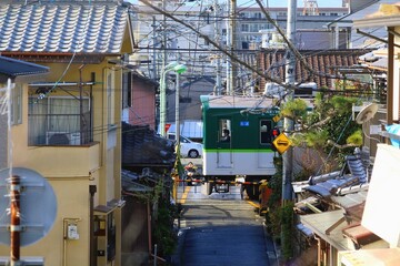 古い住宅街の路地の先にある踏切を通過する緑色の電車 © JtO