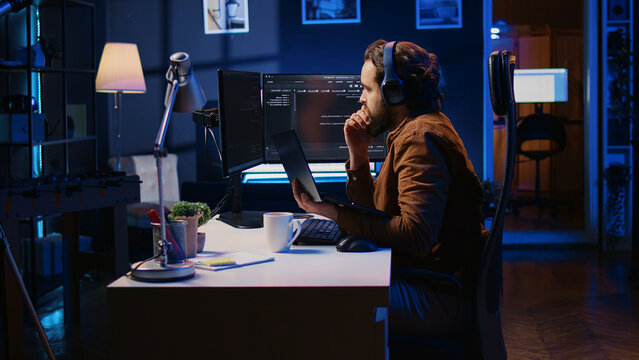 Engineer typing code in apartment office, using JavaScript programming language to develop software application, IT admin using multimonitor desktop setup and notebook to finish tasks.