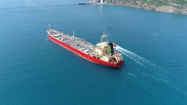 Handymax chemical tanker underway with pilot boat escort along rocky coastline, aerial view showing red hull and deck pipeline manifolds.