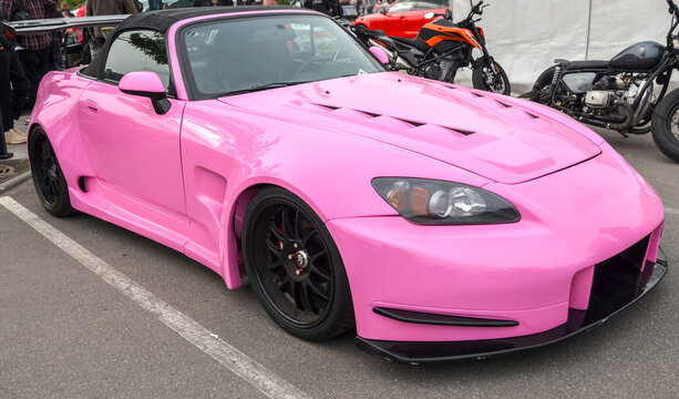 Pink Honda S2000 with widebody kit and vented hood is on display at car meet. This JDM icon features black soft top and aggressive aero, standing out among the show cars