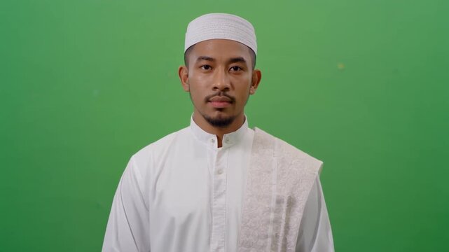 Portrait of a young Muslim man wearing a traditional white cap and robe against a green screen background.