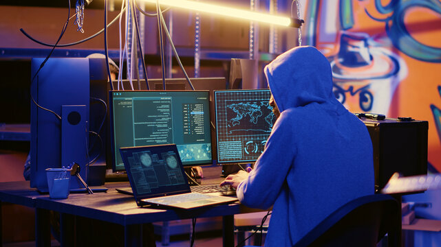 Hacker in abandoned warehouse with graffiti walls writing code that spoofs his location, tricking cops, rogue developer running script on many devices pinging wrong location to cybercriminal police.