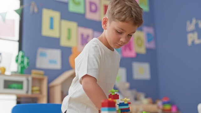 Blonde boy stacking colorful plastic blocks with hands on table in classroom; concentration learning play.