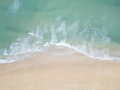 Aerial view of turquoise ocean waves crashing onto a pristine sandy beach with white foam and clear water in Padstow, Cornwall, United Kingdom.