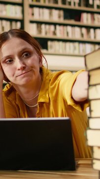 Vertical format shot: Worried young woman studying materials on laptop at library table, between tall stacks at library, Student showing academic stress and exam preparation workload pressure.
