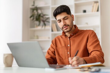 Fototapeta premium Focused Young Hispanic Man Working Diligently On Laptop While Taking Notes At Home Office, Surrounded By Shelves And Plants, Concentrated On Task. Professional Business Concept.