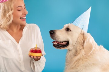 Obraz premium Smiling Young Caucasian Woman Celebrating Dog's Birthday By Holding Cake With Candle, Happy Atmosphere In Studio Setting, Joyful Moment With Pet. Family And Pet Celebration Concept.