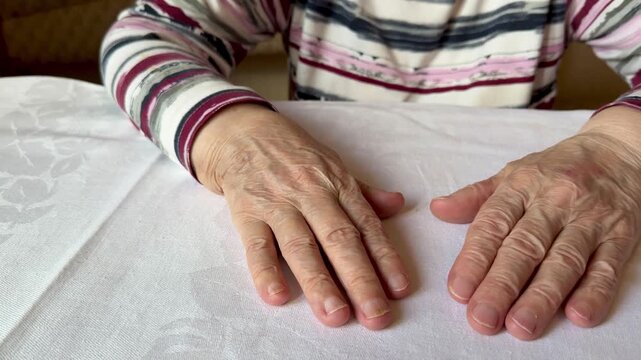 Close view of an elderly woman's hands and forearms resting palm-down on a white textured tablecloth. Striped long-sleeve shirt sleeves frame visible veins.