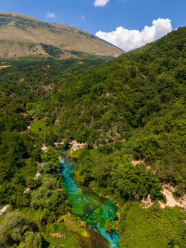 Aerial view of the bright turquoise Blue Eye water spring surrounded by dense green forest and mountain slopes in Finiq, Vlore County, Albania.