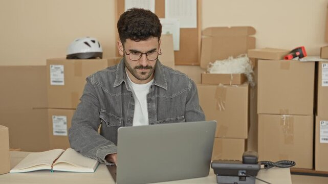 Man typing on laptop at packing desk amid cardboard boxes in warehouse building; stress perseverance.