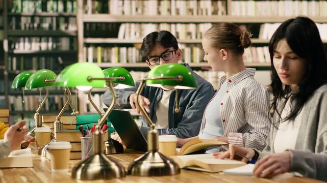 Young university students read and write at shared library table with books and green lamps.