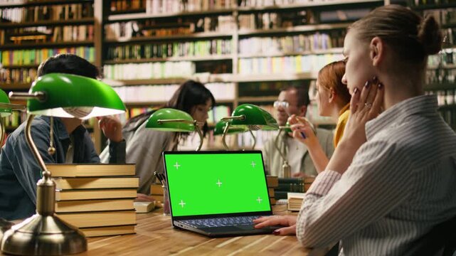Green screen chroma key on laptop at library wooden desk, study space framed by blurred students and green lamps.