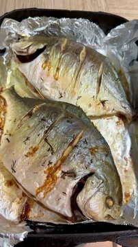 Two golden pompano on black platter, oven roasted on foil with scored skin, seasoned with salt and pepper, lemon slices tucked.