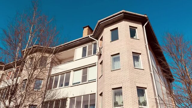 White brick apartment against blue sky. Sunlit multistorey facade with rows of windows, balconies and chimney framed by bare.