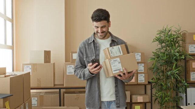 Man holding stacked parcels and smartphone, smiling while checking messages and balancing boxes in a building; entrepreneurial pride.