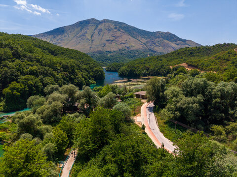 Aerial view of the Blue Eye natural spring surrounded by dense green forest and a winding road with a large mountain in the background in Finiq, Vlore County, Albania.