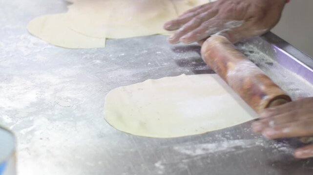 close-up view of hands using a rolling pin to flatten dough on a floured surface, shows a kitchen preparation process with thin dough pieces being shaped before cooking.