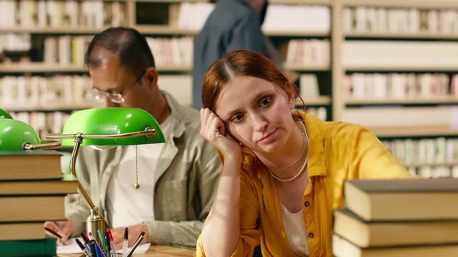 Frustrated young woman frowning at laptop between tall book stacks at library desk, showing academic pressure during long study session and coursework, Burnout challenge for research and exam stress.