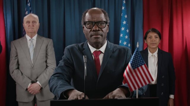 Portrait of Black senior politician standing at podium with US flag, flanked by diverse colleagues. Video is suitable for political campaigns and government communications