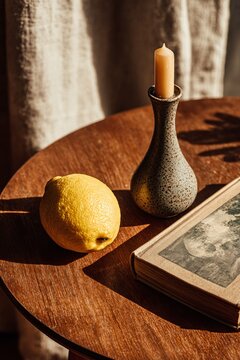 Mid-century modern still life flat-lay on round teak table with whole lemon, lit taper candle in sculptural ceramic holder, vintage graphic book, warm natural window light and subtle film grain.