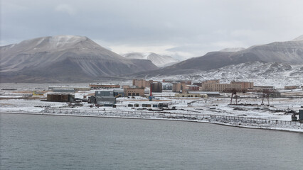 Aerial winter view of a remote arctic industrial port with cranes and warehouses nestled between snowy mountains and calm, icy waters under a heavy overcast sky. © Lihui