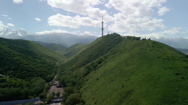 Aerial view of Kok Tobe television tower on a green hill with snow-capped mountains in Almaty, Kazakhstan.