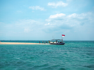 Obraz premium A small boat with an Indonesian flag sails in the turquoise waters near a tropical island sandbar, where a group of people are gathered.