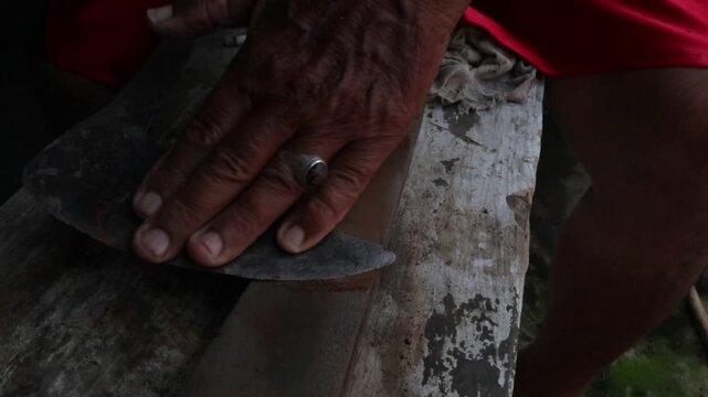 A man sharpens a machete manually with a sharpening stone in a rural village environment. Close-up footage showing traditional tool maintenance, agricultural preparation, and skilled handwork.
