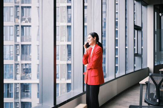 Hispanic businesswoman, female CEO in casual red suit, talking on mobile phone in modern workplace, confident job discussing communication near office window view, city building in urban lifestyles.
