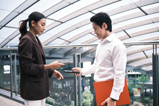 Young Asian smart businesspeople exchange digital contact information with NFC QR code, wireless mobile phone app technology, contactless startup collaboration at outdoor workplace, modern lifestyle.