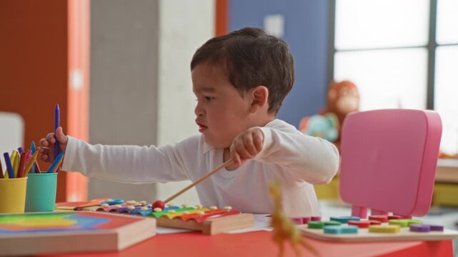 Boy striking a colorful xylophone with a mallet and reaching for pencils on a red table in a building; curiosity learning.