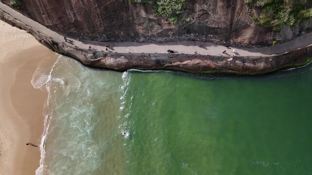 Aerial view of Praia do Leme with people walking along the coastal path by rocky cliffs and turquoise ocean water in Rio de Janeiro, State of Rio de Janeiro, Brazil.